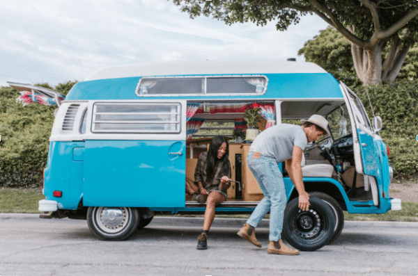 Person 1 rolling spare tire in front of van. Person two sitting and watching person 1. The man will be able to easily change his tire using the 9 steps outlined in the article